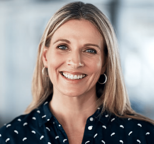 Professional woman smiling at camera, wearing patterned dark blouse, indoor office setting.