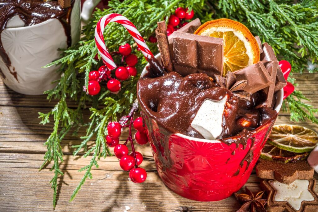 Festive hot chocolate with chocolate pieces and marshmallow in red mug surrounded by holiday decorations.