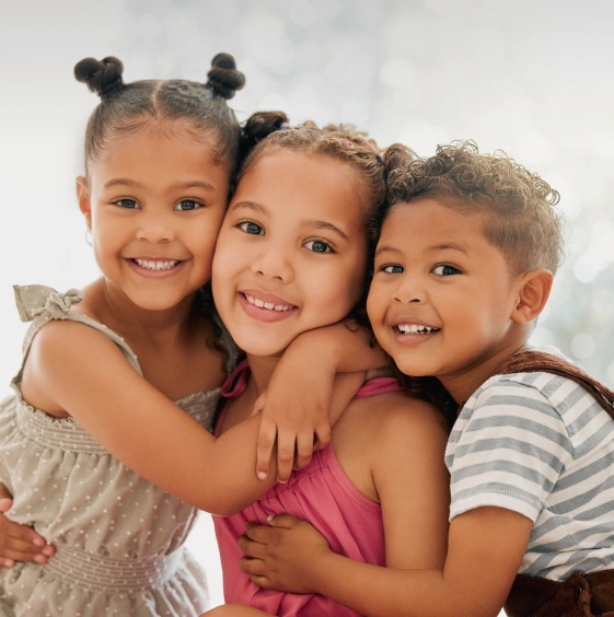 A close-up studio portrait of three smiling, young Black children embracing each other tightly.