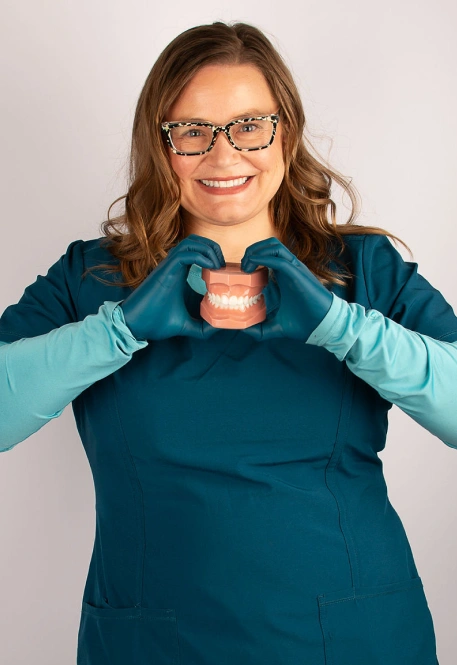 Dentist in glasses making a heart shape with gloved hands around model teeth
